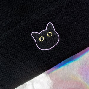 Op de afbeelding: Zwarte beanie met een wit geborduurd kattengezicht met gele ogen.