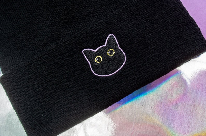 Op de afbeelding: Zwarte beanie met een wit geborduurd kattengezicht met gele ogen.