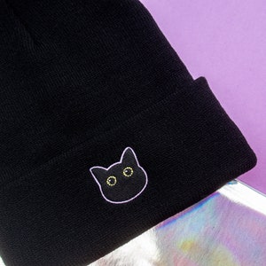 Op de afbeelding: Zwarte beanie met een zwarte kat geborduurd op de voorkant. De kat heeft gele ogen en een witte omtrek.