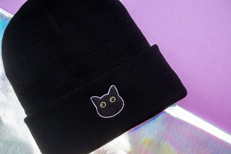 Op de afbeelding: Zwarte beanie met een zwarte kat geborduurd op de voorkant. De kat heeft gele ogen en een witte omtrek.