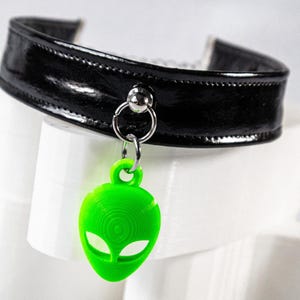 Op de afbeelding: Een zwarte, glanzende choker-stijl kraag met een zilveren ring en een felgroene alien hanger. De alien hanger heeft grote ogen en een gestructureerd oppervlak. De kraag is gemaakt van een glanzend materiaal.