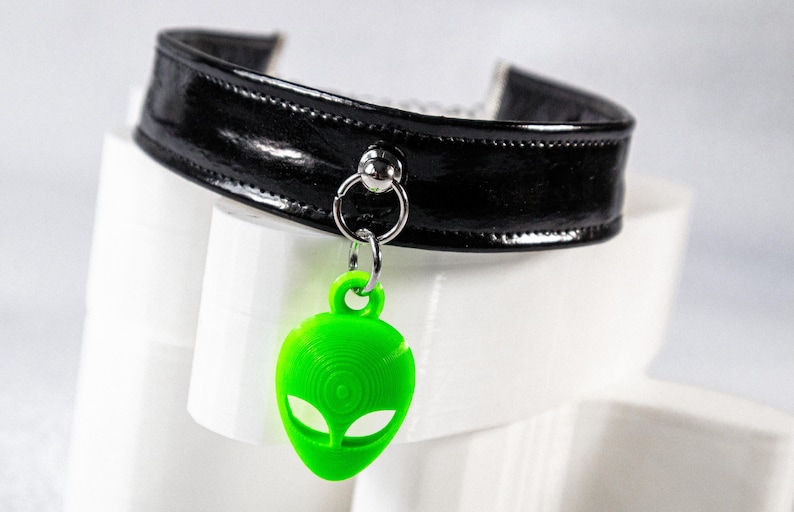 Op de afbeelding: Een zwarte, glanzende choker-stijl kraag met een zilveren ring en een felgroene alien hanger. De alien hanger heeft grote ogen en een gestructureerd oppervlak. De kraag is gemaakt van een glanzend materiaal.