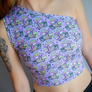 May include: A lavender one-shoulder crop top featuring a repeating pattern of fast food items with skull accents. The design includes burgers, fries, and drinks. The top is made of a soft, stretchy material.