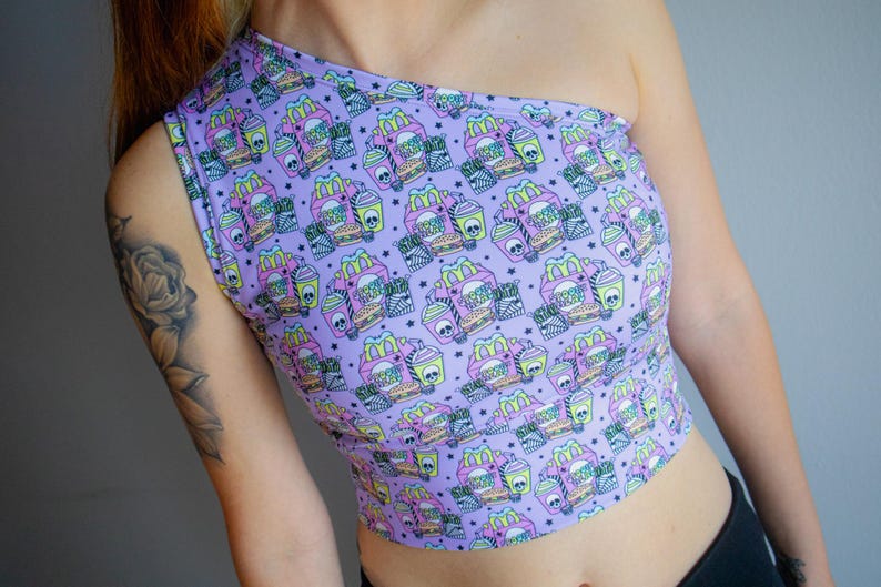May include: A lavender one-shoulder crop top featuring a repeating pattern of fast food items with skull accents. The design includes burgers, fries, and drinks. The top is made of a soft, stretchy material.
