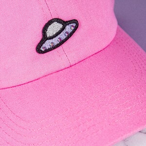 以下が含まれることがあります： ラインストーンが付いた黒と白の刺繍されたUFOパッチが付いたピンクの野球帽