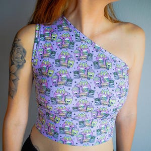 May include: A lavender one-shoulder crop top with a repeating pattern of fast food items, including burgers, fries, and drinks, with skull and star accents. The top is made of a soft, stretchy material.