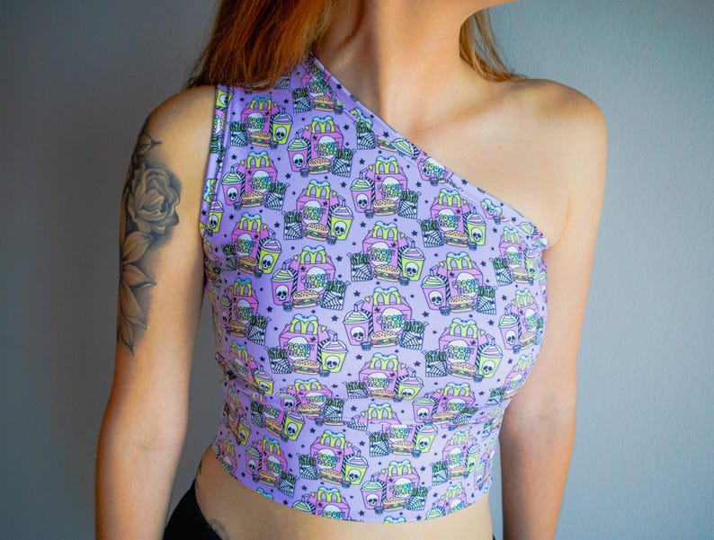 May include: A lavender one-shoulder crop top with a repeating pattern of fast food items, including burgers, fries, and drinks, with skull and star accents. The top is made of a soft, stretchy material.