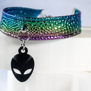 Op de afbeelding: Een choker ketting met een getextureerde, iriserende afwerking in tinten blauw, groen en paars. Een zwarte alien hoofd hanger hangt in het midden. De choker wordt gepresenteerd op een wit oppervlak.