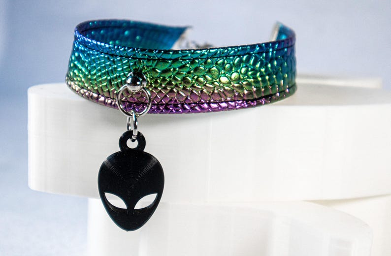 Op de afbeelding: Een choker ketting met een getextureerde, iriserende afwerking in tinten blauw, groen en paars. Een zwarte alien hoofd hanger hangt in het midden. De choker wordt gepresenteerd op een wit oppervlak.