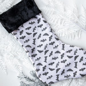 May include: A white Christmas stocking with a black faux fur cuff and a repeating pattern of black bats.