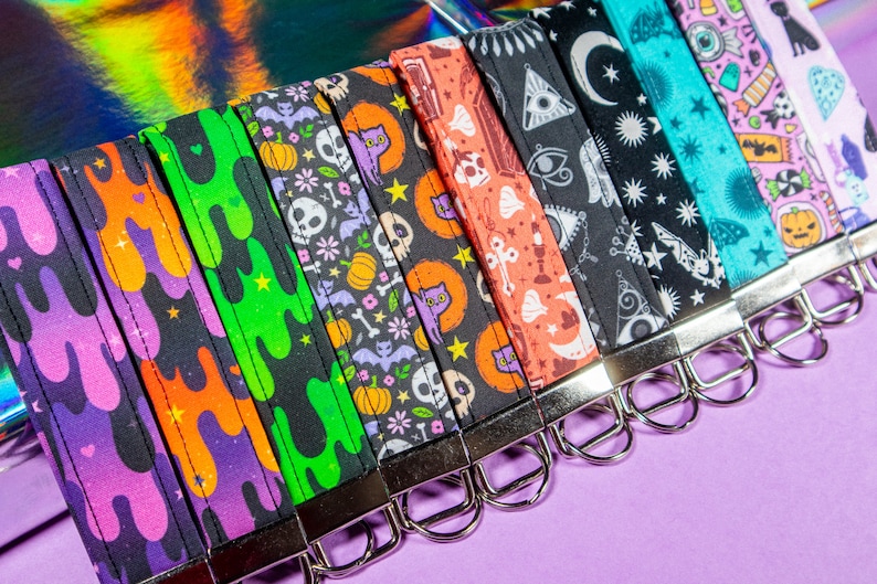 May include: A set of nine colorful keychains with different spooky and whimsical patterns. The keychains feature designs like dripping slime, skulls, bats, pumpkins, and stars. The keychains are all black with colorful accents.