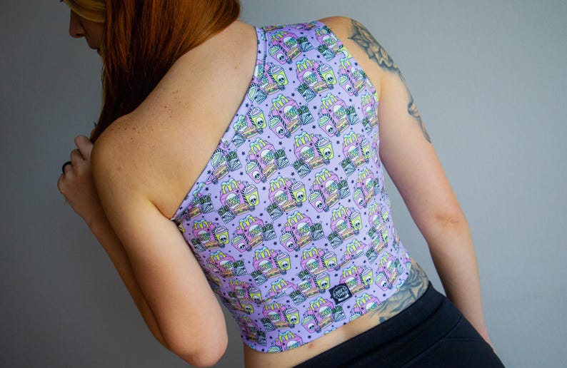 May include: A lavender crop top with a repeating pattern of cartoon fast food items in pastel colors. The top has a single shoulder strap and a small tag at the bottom.
