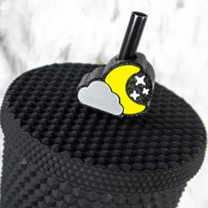 May include: A black tumbler with a textured surface, topped with a straw and a charm. The charm features a yellow crescent moon, white stars, and a gray cloud design, creating a night sky theme.