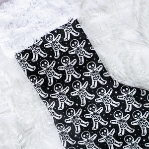 Puede incluir: Una media de Navidad negra con un puño de piel sintética blanca. La media presenta un patrón repetido de hombres de jengibre esqueleto blancos sobre un fondo negro con estrellas blancas.