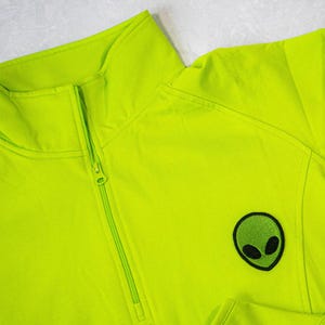 Op de afbeelding: Een felgroen ritsvest met een geborduurd groen alien gezicht op de rechtermouw.