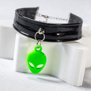 Op de afbeelding: Zwarte lakleren choker ketting met een zilveren ketting en een felgroene alien hanger. De alien hanger heeft een gedetailleerd ontwerp en hangt aan een zilveren ring. De choker wordt gepresenteerd op een witte ondergrond.