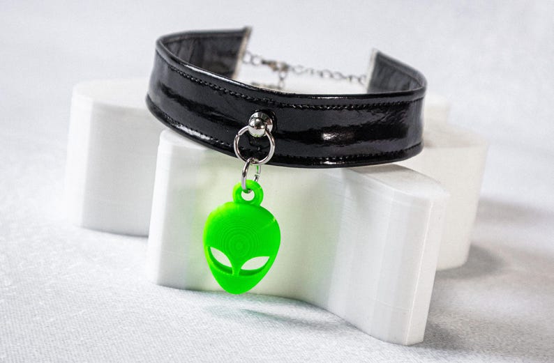 Op de afbeelding: Zwarte lakleren choker ketting met een zilveren ketting en een felgroene alien hanger. De alien hanger heeft een gedetailleerd ontwerp en hangt aan een zilveren ring. De choker wordt gepresenteerd op een witte ondergrond.