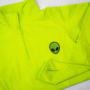 Op de afbeelding: Een felgroen, neonkleurig, 1/4-rits trui met een klein groen alien-motief op de linkerborst.