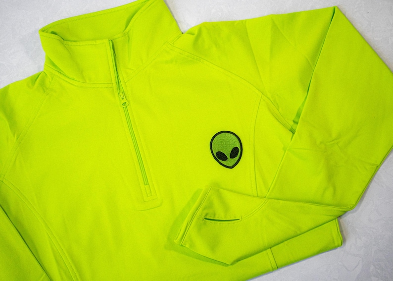 Op de afbeelding: Een felgroen, neonkleurig, 1/4-rits trui met een klein groen alien-motief op de linkerborst.