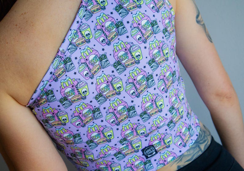 May include: A lavender halter top with a repeating pattern of fast food items and skulls. The design includes burgers, fries, and drinks in pink, yellow, and white. The top has a small black tag with white text.