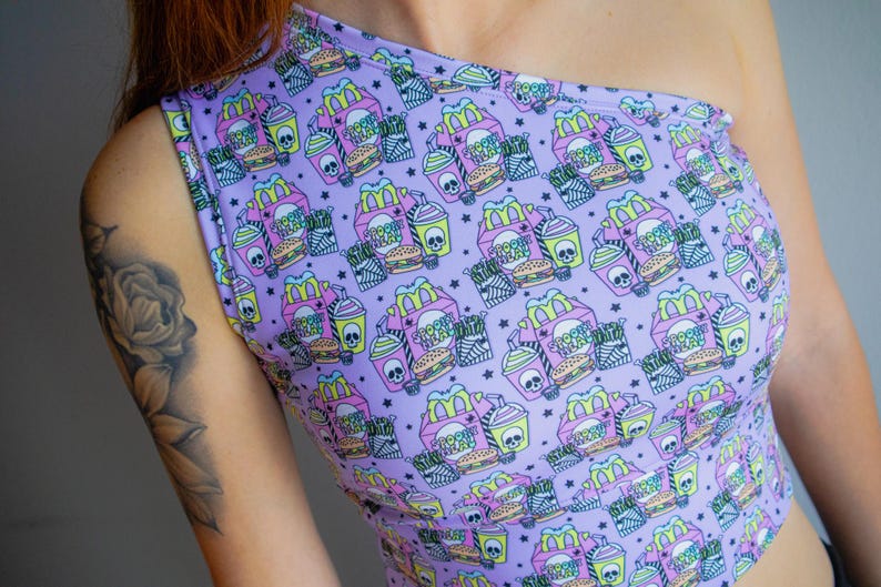 May include: A lavender one-shoulder crop top with a repeating pattern of fast food boxes, burgers, fries, and drinks, each with a skull design. The text "SPOOKY TREAT" is visible on the boxes. The top is made of a soft, stretchy material.