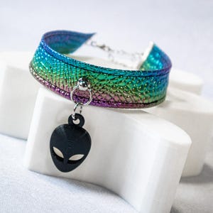 Op de afbeelding: Een choker-ketting met een regenboogkleurige iriserende afwerking. De choker heeft een zwarte alien-hoofdhanger. De choker wordt gepresenteerd op een wit oppervlak.