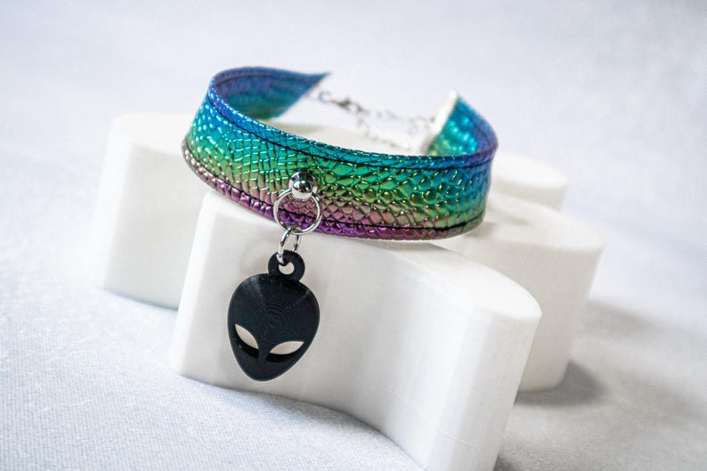 Op de afbeelding: Een choker-ketting met een regenboogkleurige iriserende afwerking. De choker heeft een zwarte alien-hoofdhanger. De choker wordt gepresenteerd op een wit oppervlak.