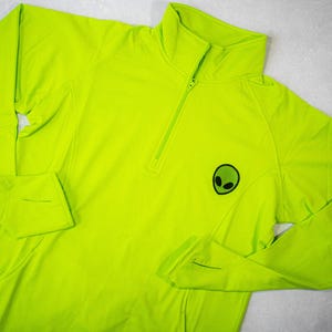 Op de afbeelding: Een fel neon groen shirt met lange mouwen, een halve rits en een klein alien-motief op de voorkant.