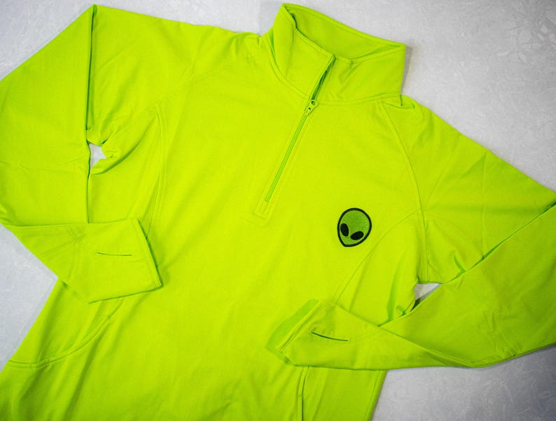 Op de afbeelding: Een fel neon groen shirt met lange mouwen, een halve rits en een klein alien-motief op de voorkant.