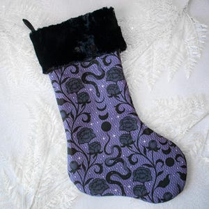 May include: A Christmas stocking with a black faux fur cuff and a purple body. The body of the stocking features a floral and snake pattern in black. The background is a white, textured surface.