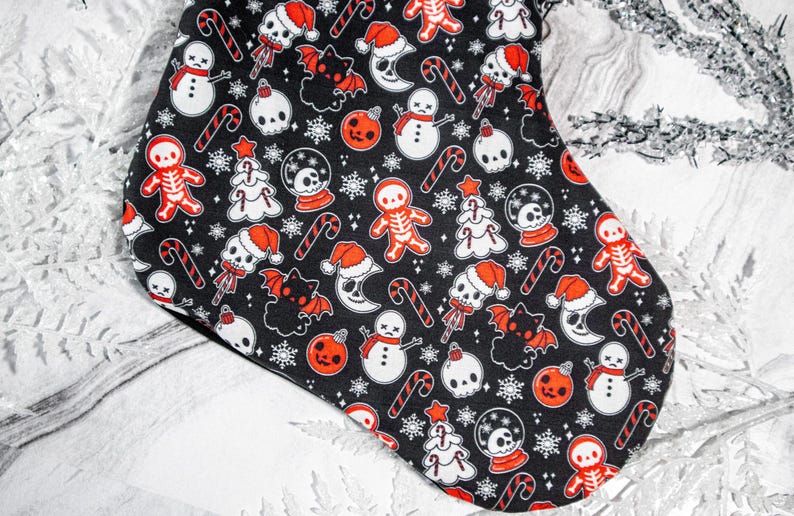 Puede incluir: Una media de Navidad negra con un patr&oacute;n blanco y rojo de calaveras, mu&ntilde;ecos de nieve, bastones de caramelo, murci&eacute;lagos y hombres de jengibre. La media est&aacute; decorada con un tema navide&ntilde;o espeluznante.