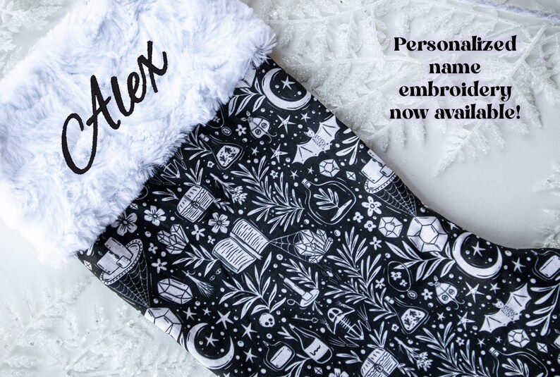 May include: A black and white Christmas stocking with a white faux fur cuff. The stocking features a repeating pattern of moons, stars, crystals, and other witchy imagery. The name "Alex" is embroidered on the cuff in black thread.