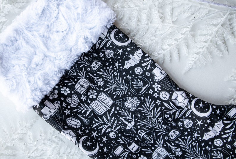 May include: A black Christmas stocking with a white faux fur cuff. The stocking features a white print of various magical and mystical objects, including moons, stars, crystals, bottles, and a bat.