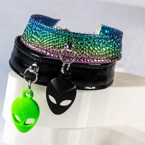 Op de afbeelding: Twee chokers met buitenaardse bedels. De ene is zwart, de andere iriserend met regenboogkleuren. De bedels zijn een groene en een zwarte alienkop. De chokers worden getoond op een witte, gelaagde sokkel.