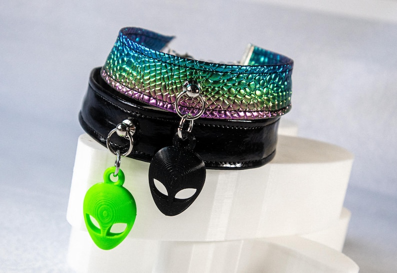 Op de afbeelding: Twee chokers met buitenaardse bedels. De ene is zwart, de andere iriserend met regenboogkleuren. De bedels zijn een groene en een zwarte alienkop. De chokers worden getoond op een witte, gelaagde sokkel.