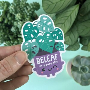 May include: A sticker featuring a cartoon plant in a purple pot with the text "BELEAF in yourself!". The plant has green leaves with white accents. The sticker has a white border and is held against a background of green plants.