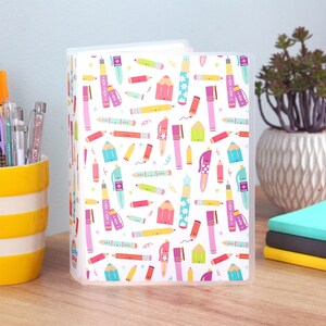 May include: A clear plastic binder with a white background covered in colorful illustrations of pens, pencils, and markers. The binder is standing upright on a wooden surface. A yellow and white striped cup with pens and pencils is to the left.