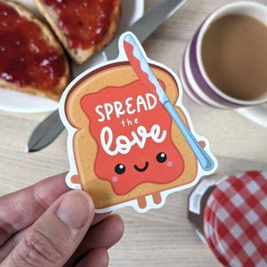 May include: A sticker depicting a cartoon slice of toast with jam and a smiling face. The text "SPREAD the love" is written on the jam. A blue knife is next to the toast. A cup of tea and toast with jam are in the background.