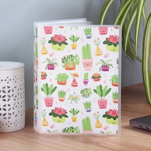 May include: A clear, plastic binder with a white cover featuring a repeating pattern of colorful potted plants. The plants include succulents, flowering plants, and hanging plants in various pots and vases. The binder is open, revealing white pages.