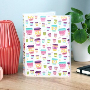 May include: A clear plastic binder with a white cover featuring a repeating pattern of colorful coffee cups. The cups have various floral and geometric designs in shades of pink, yellow, and teal. A red vase is on the left.