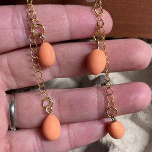 Pode incluir: Um par de brincos pendentes com um amuleto de caveira de cer&acirc;mica branca e tr&ecirc;s contas em forma de ovo cor de p&ecirc;ssego penduradas em uma corrente dourada.