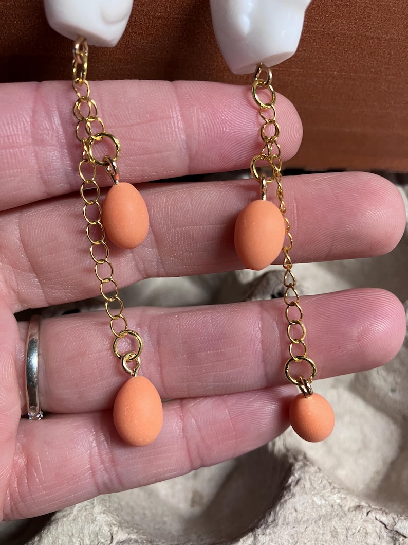 Pode incluir: Um par de brincos pendentes com um amuleto de caveira de cer&acirc;mica branca e tr&ecirc;s contas em forma de ovo cor de p&ecirc;ssego penduradas em uma corrente dourada.