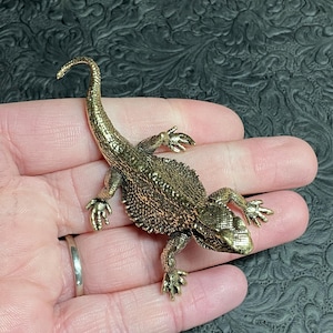May include: A detailed bronze-colored lizard figurine with textured scales and a curled tail. The reptile is posed on a hand, showcasing its intricate design. The lizard's feet are detailed, and the overall aesthetic is antique.