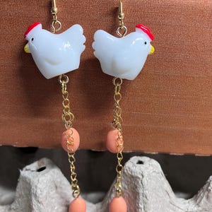 Pode incluir: Um par de brincos de ouro pendurados com pingentes de frango de cer&acirc;mica branca, cristas vermelhas e detalhes de contas cor de p&ecirc;ssego.