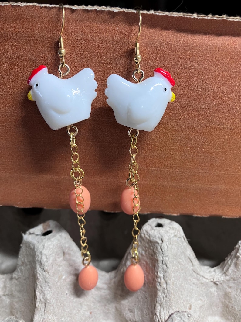 Pode incluir: Um par de brincos de ouro pendurados com pingentes de frango de cer&acirc;mica branca, cristas vermelhas e detalhes de contas cor de p&ecirc;ssego.