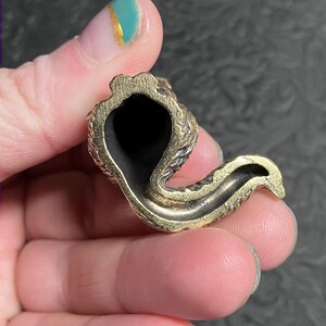 May include: A small, brass-colored pipe with a curved design. The pipe has a dark interior and a textured exterior. The object is held in a hand.