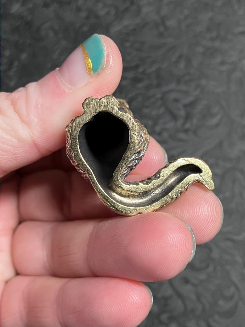May include: A small, brass-colored pipe with a curved design. The pipe has a dark interior and a textured exterior. The object is held in a hand.