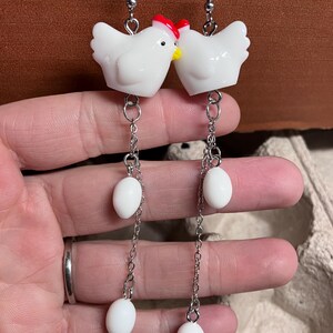 Pode incluir: Brincos de cer&acirc;mica branca em forma de frango com cristas vermelhas e pingentes de ovo branco em correntes prateadas.