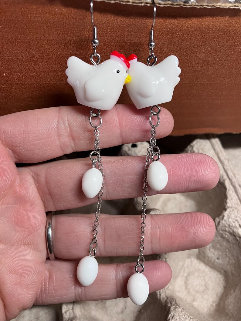 Pode incluir: Brincos de cer&acirc;mica branca em forma de frango com cristas vermelhas e pingentes de ovo branco em correntes prateadas.