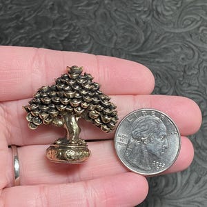 May include: A small, bronze-colored tree sculpture with a detailed design. The tree has a rounded top and a detailed trunk and base. A U.S. quarter coin is next to the tree for size comparison.